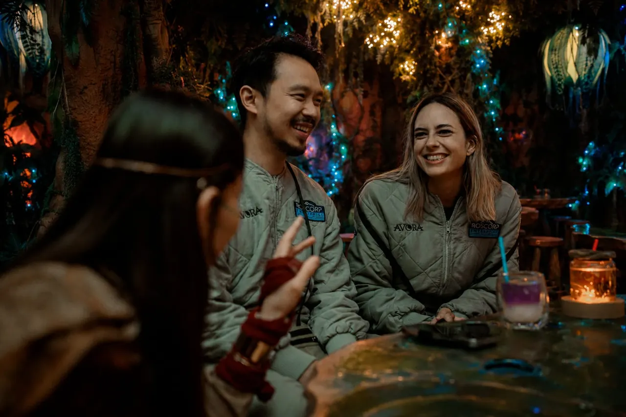 Three young adults in uniform jackets laugh together in an illuminated cave environment decorated with glowing blue and golden lights strung through trees.