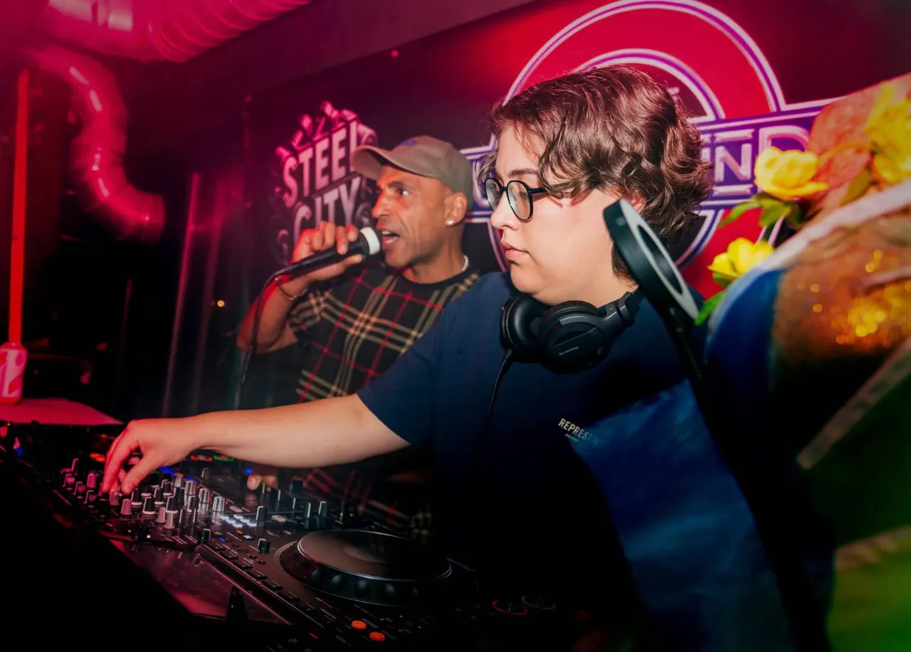 DJ operating turntables at a nightclub whilst a performer sings into a microphone, illuminated by red neon lighting.