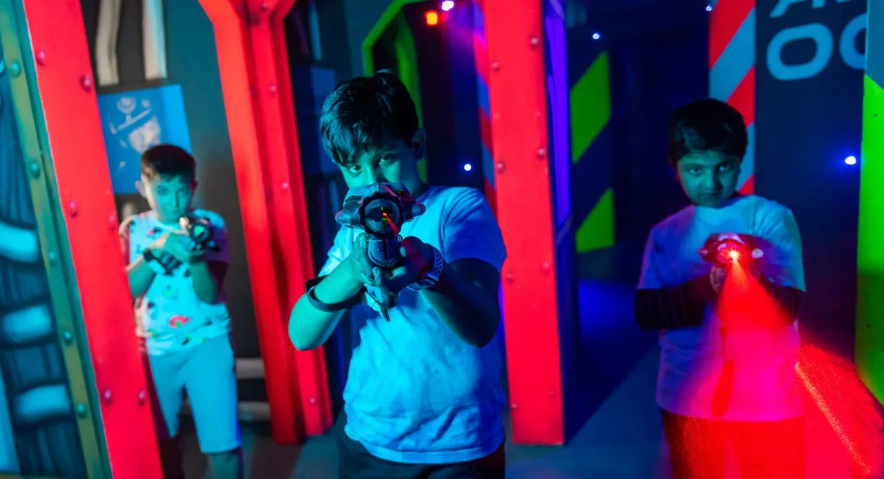 Children playing laser tag in a brightly lit arena with red, blue and green neon panels.