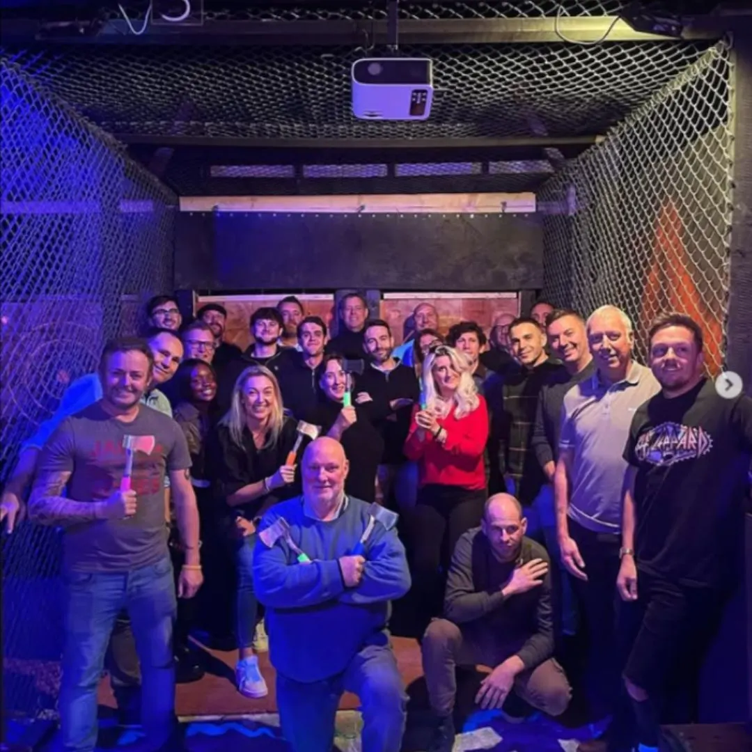 Large group of people posing together in a blue and orange lit axe throwing venue with netting on the walls.