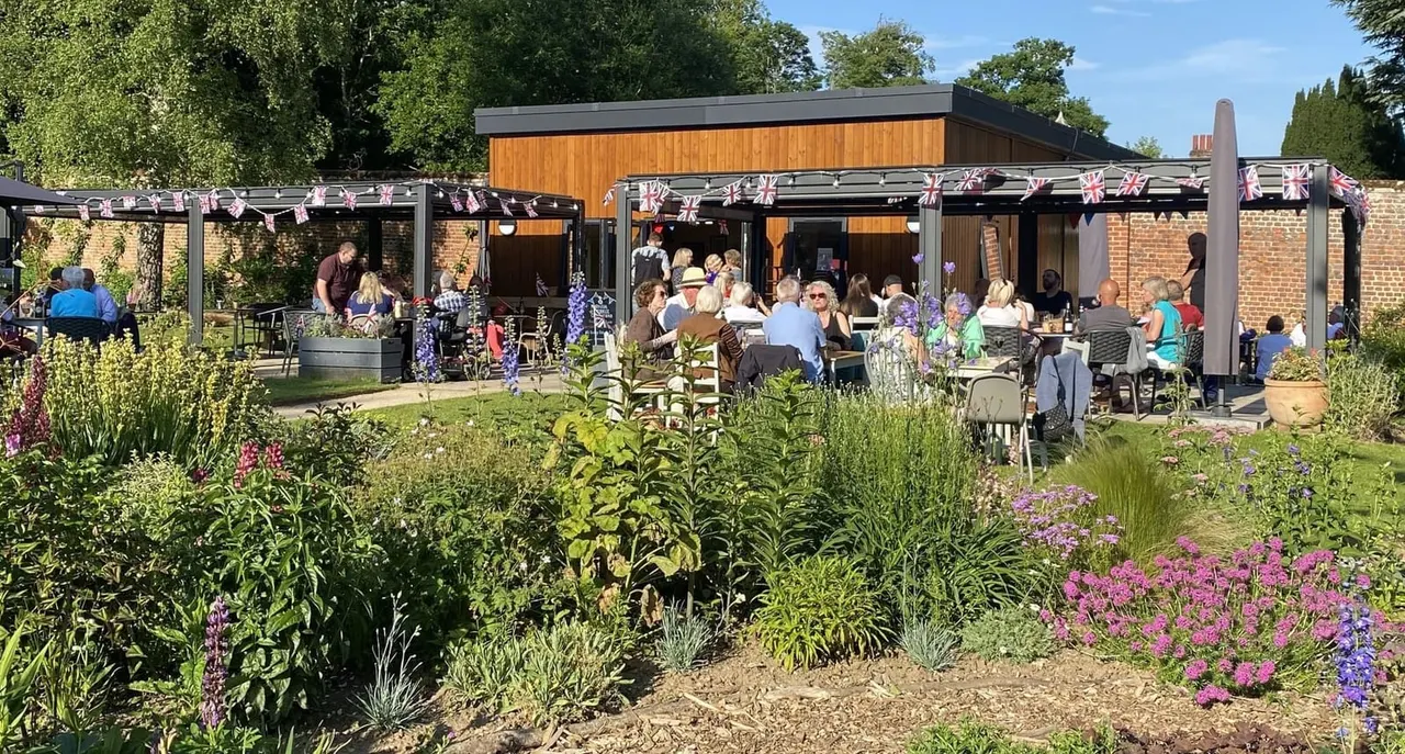 Outdoor garden café with many visitors seated at tables under pergolas decorated with Union Jack bunting, surrounded by flowering plants and shrubs.