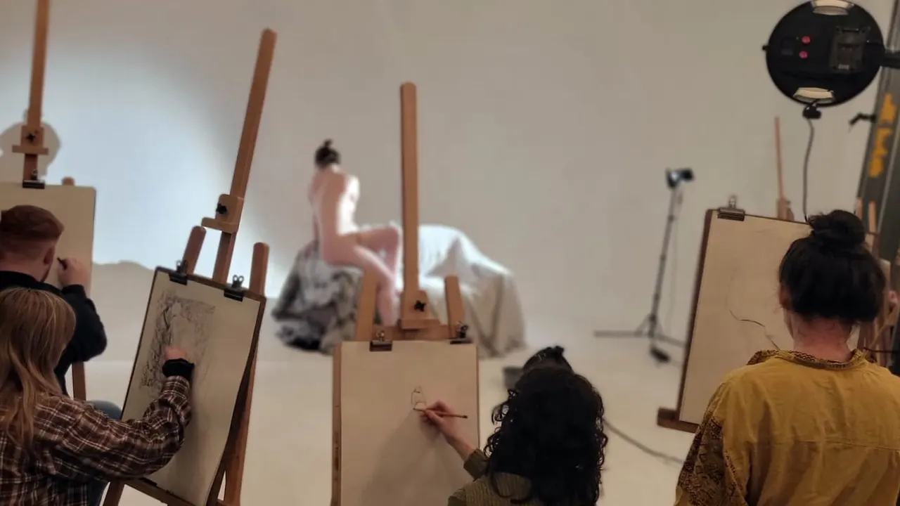 Life drawing class with multiple participants seated at easels sketching a posed model in the centre of the studio.
