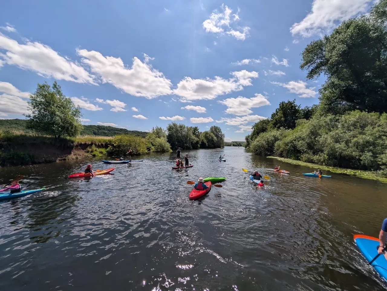 Multiple people kayaking and paddleboarding on a river surrounded by green banks and trees under blue sky.