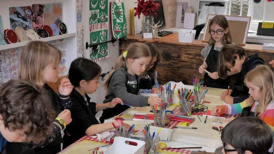 Children engaged in a creative craft activity at a table surrounded by coloured markers and art supplies in a bright studio space.