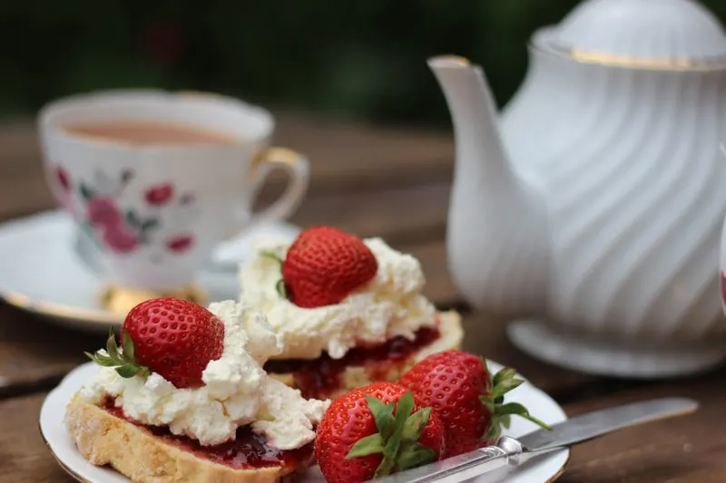 Cream tea with scones topped with jam and strawberries, alongside a teapot and floral teacup.