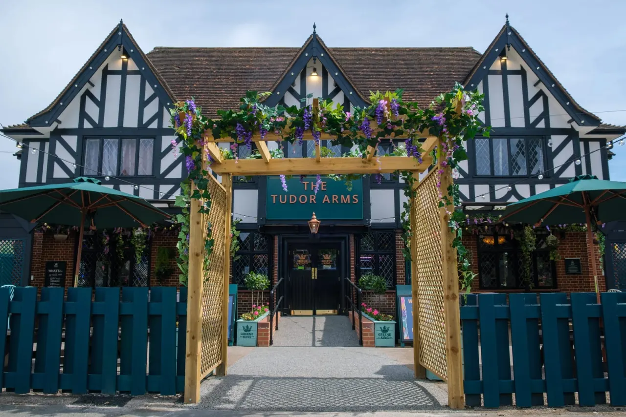 Tudor-style pub with black and white timber framing, decorated with purple wisteria hanging from a wooden pergola entrance, blue umbrellas and fencing visible to sides.