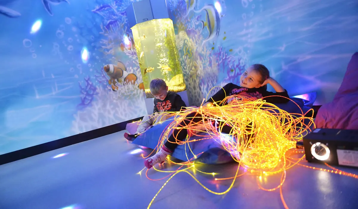 Two young children interact with illuminated interactive displays in a blue-lit immersive play environment with glowing neon elements.