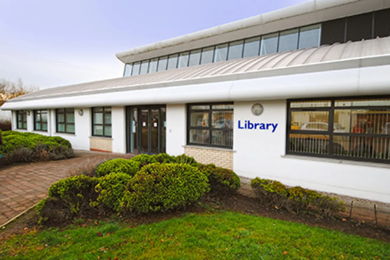 Modern single-storey white building with "Library" signage, brick paved forecourt and landscaped shrubs.