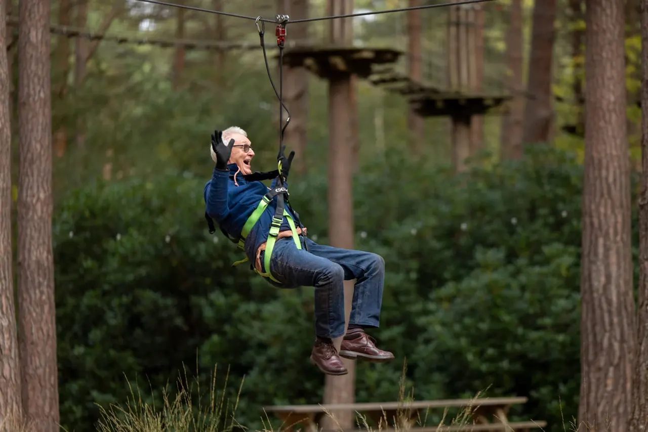 Older adult ziplining through a forest canopy on a Go Ape course, suspended from a cable between tall trees.