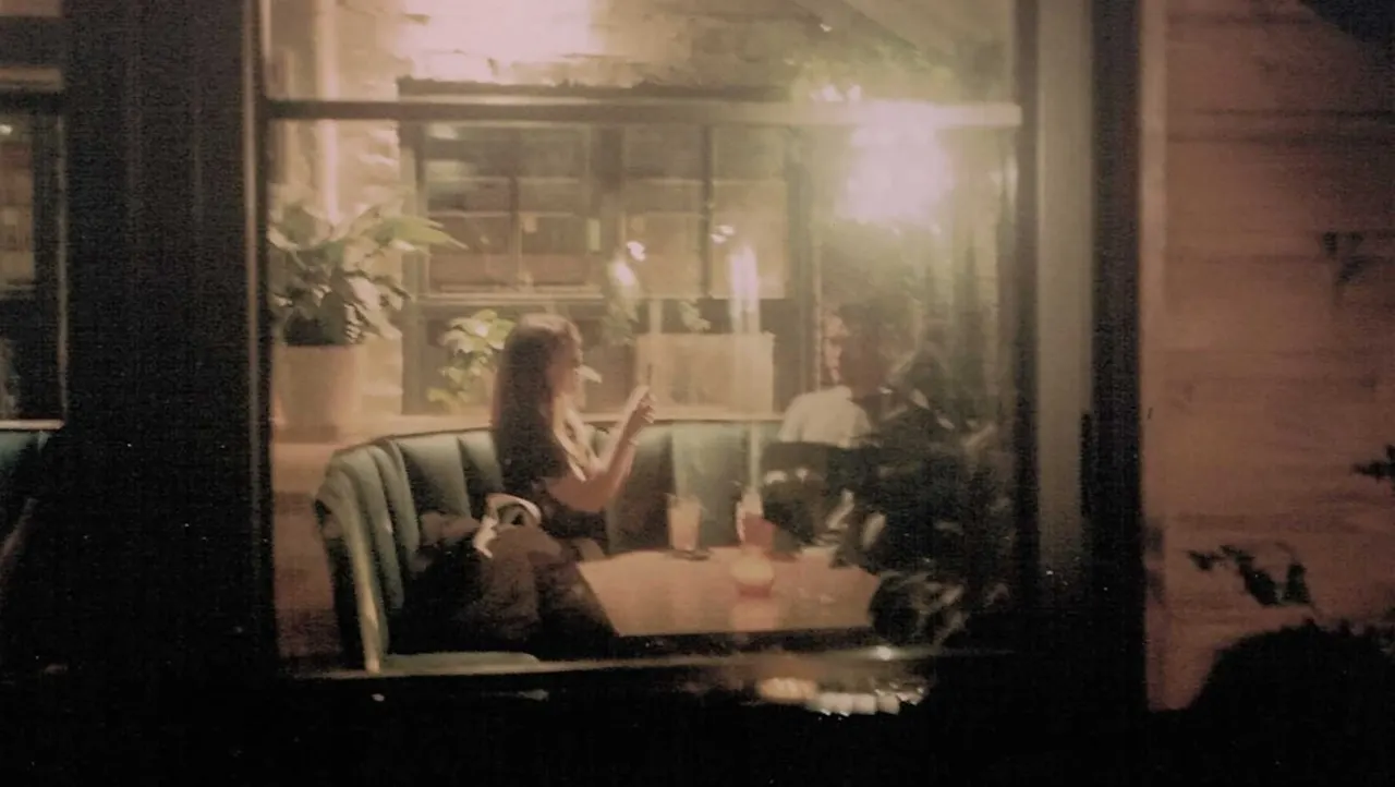 People dining together at a table inside a warmly lit restaurant or café viewed through a window at night.