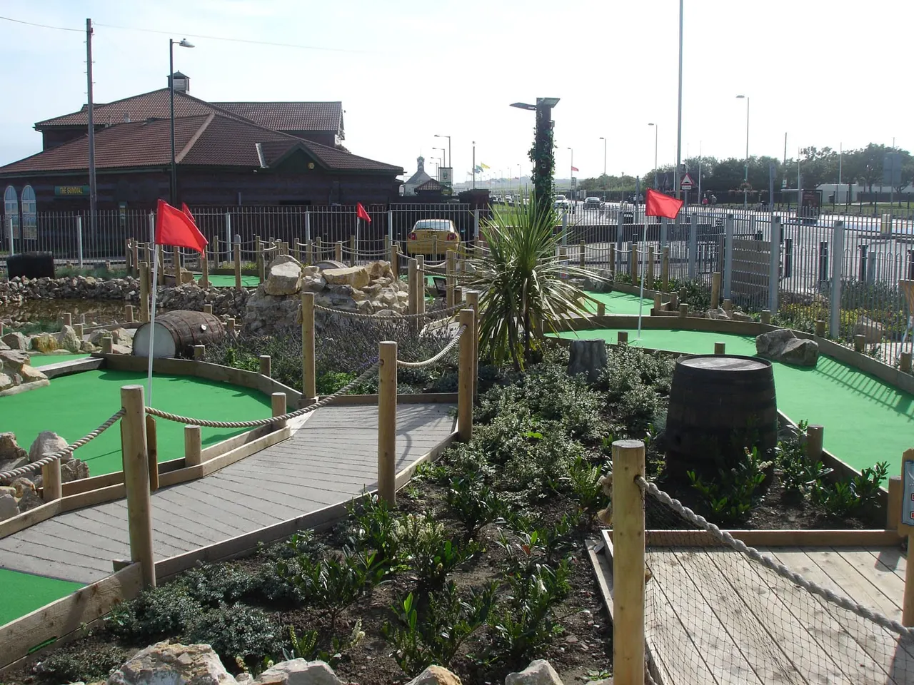 Themed mini-golf course with bright green playing surfaces, wooden bridges, red flags, and nautical décor including barrels and tropical plants.