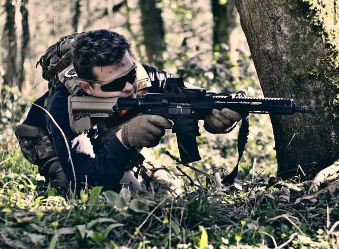 Person in tactical gear aiming a paintball marker in a woodland setting during gameplay.