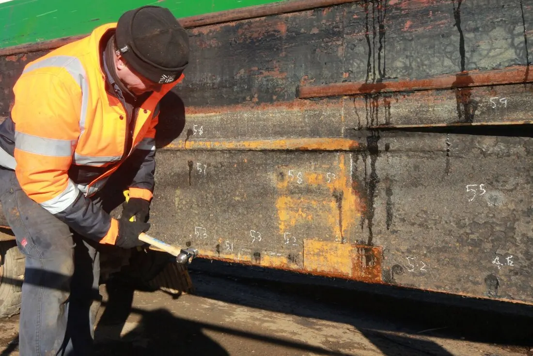 Worker in high-visibility jacket chiselling rust from the hull of a narrowboat during restoration work.