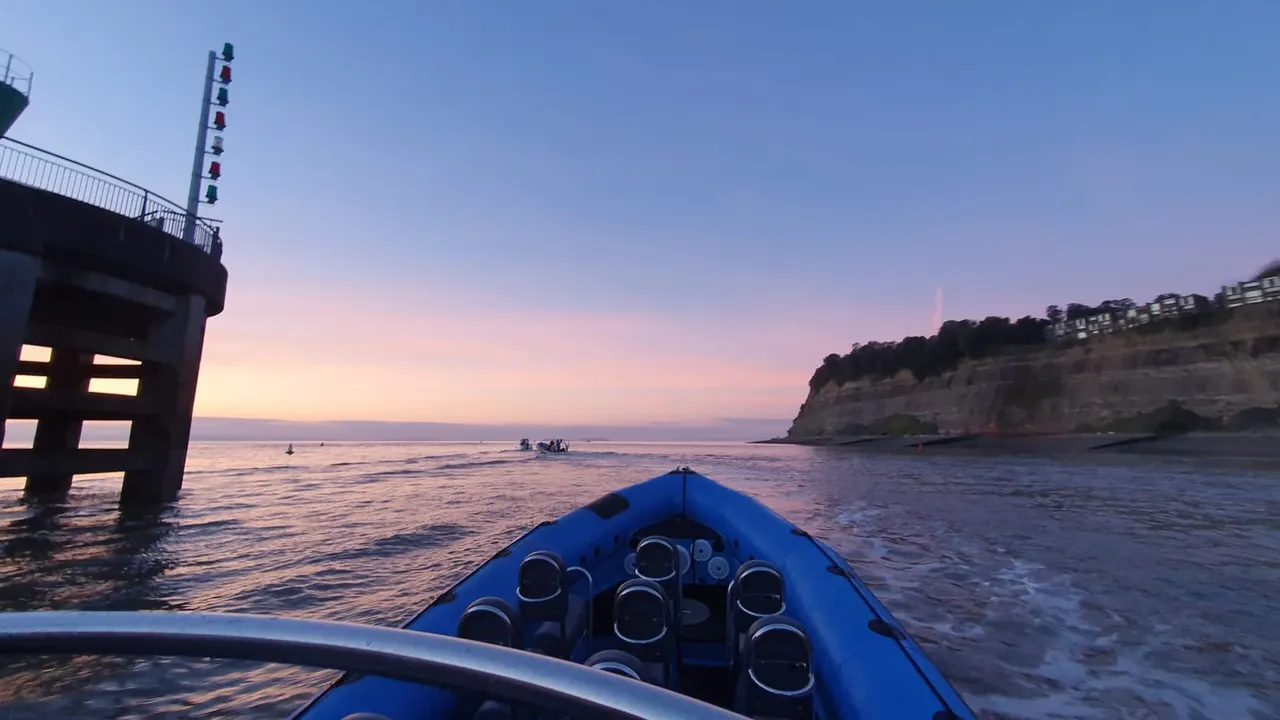 Blue speedboat with multiple engines heading out to sea at dusk, with a pier and coastal cliffs visible.