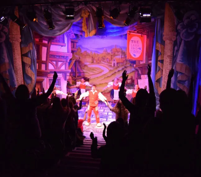 Audience members with raised arms watch colourful stage production with performers under theatrical lighting and painted backdrops.