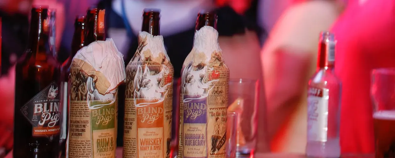 Bottles of Blind Pig branded alcoholic beverages arranged on a bar counter with pink and red ambient lighting.