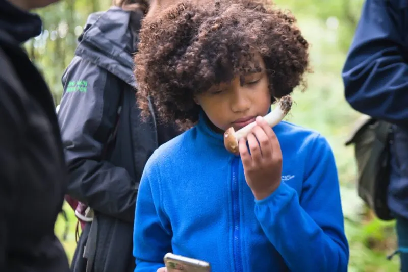 Child in blue jacket eating a snack outdoors surrounded by adults on a woodland walk.
