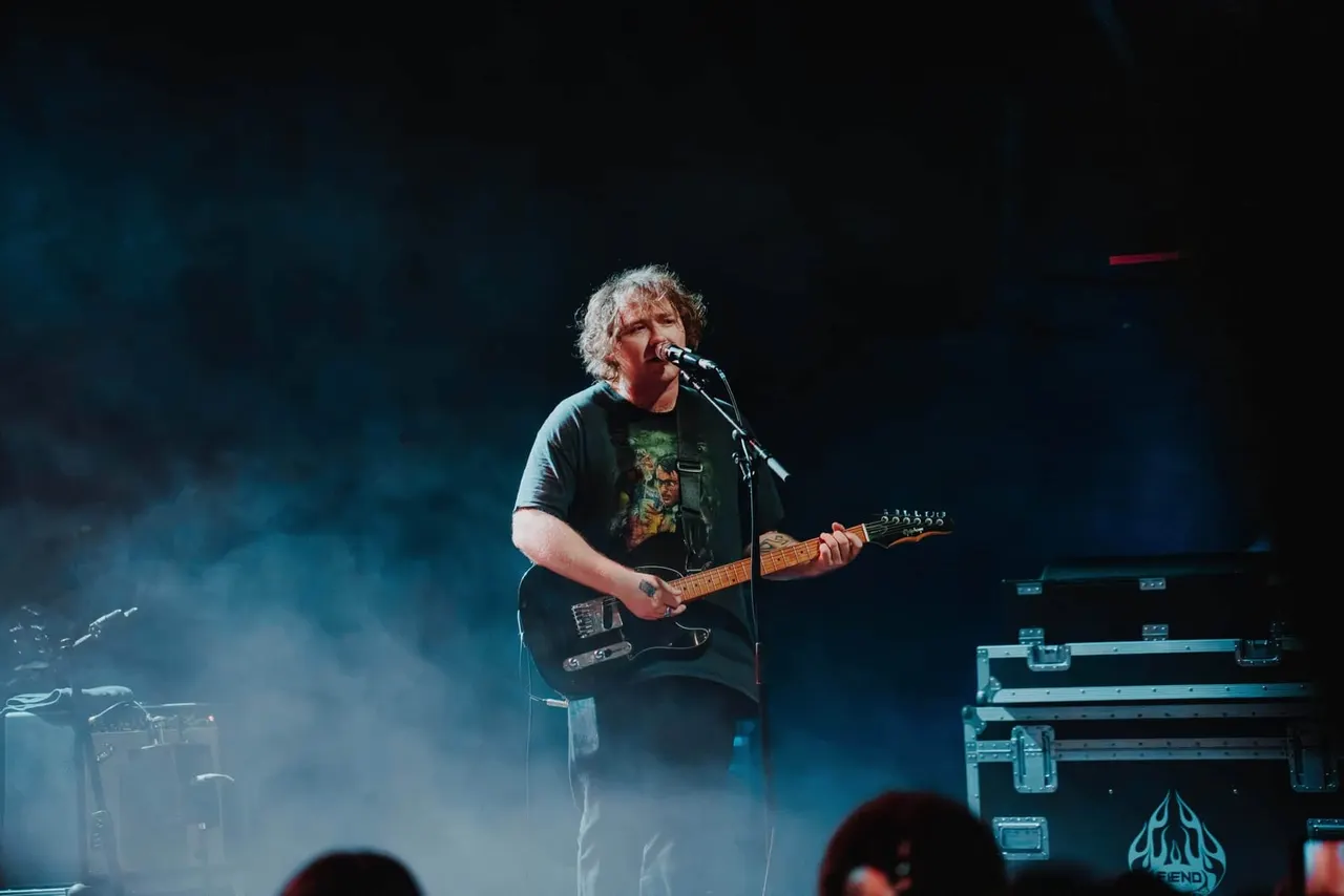 Musician with white curly hair singing and playing electric guitar at a concert venue with blue stage lighting and fog effects.