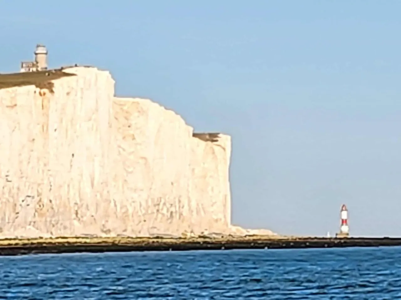 Tall white chalk cliffs with a lighthouse on top, viewed from the sea on a clear day.