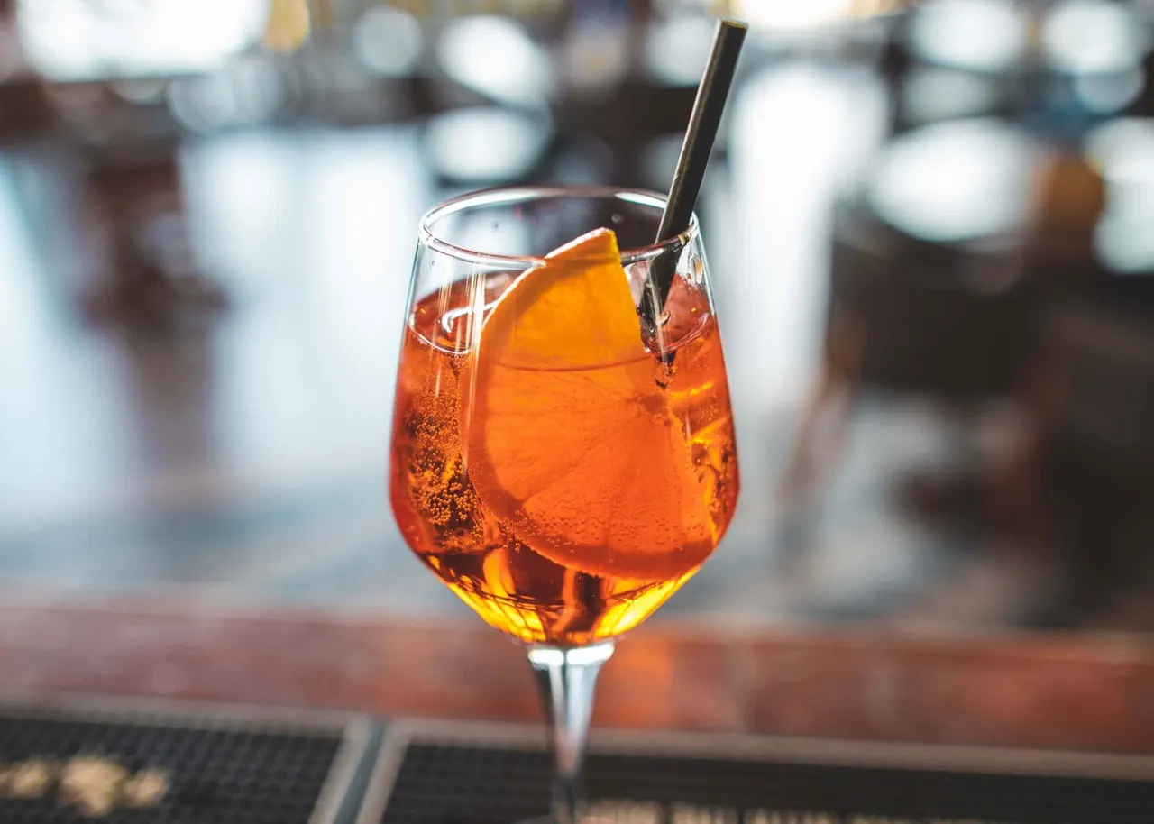 Amber-coloured cocktail with ice and orange slice garnish, served with a black straw at a bar.
