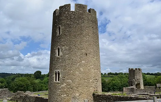 Medieval stone castle tower with crenellated top, narrow window openings, and ruins of surrounding structures under partly cloudy sky.