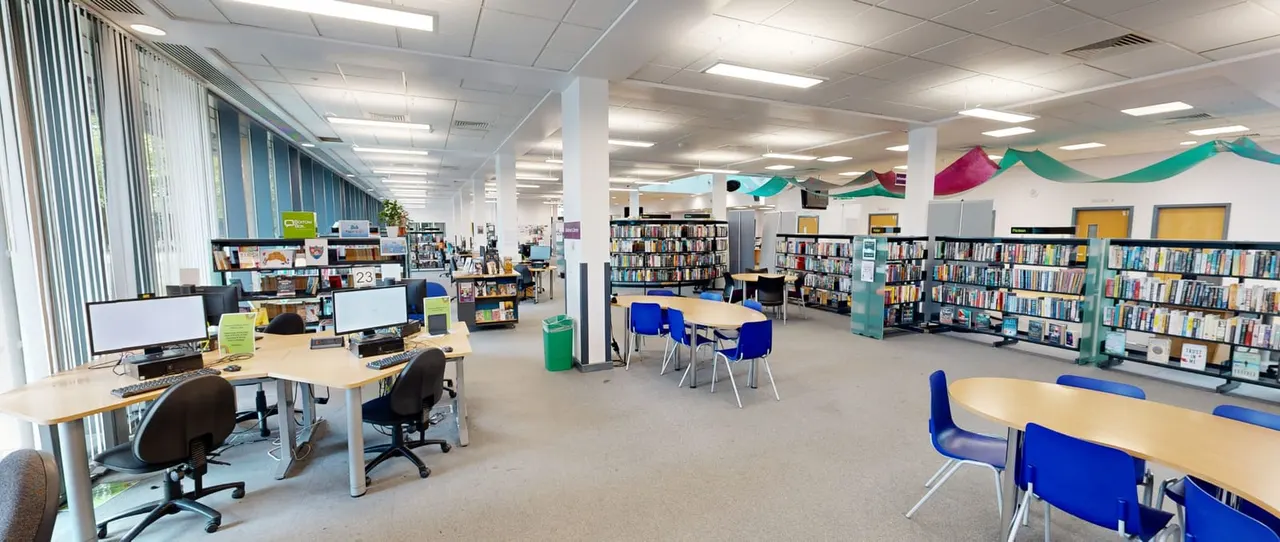 Interior of a modern library with computer workstations, bookshelves, tables with blue chairs and coloured fabric bunting suspended from ceiling.
