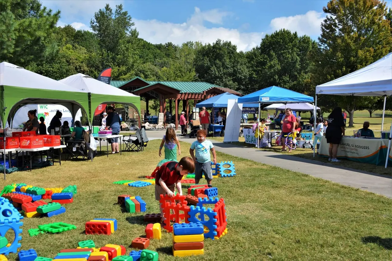 Outdoor community event with children playing on colourful foam blocks on grass, surrounded by market stalls and gazebos, trees and buildings in background.