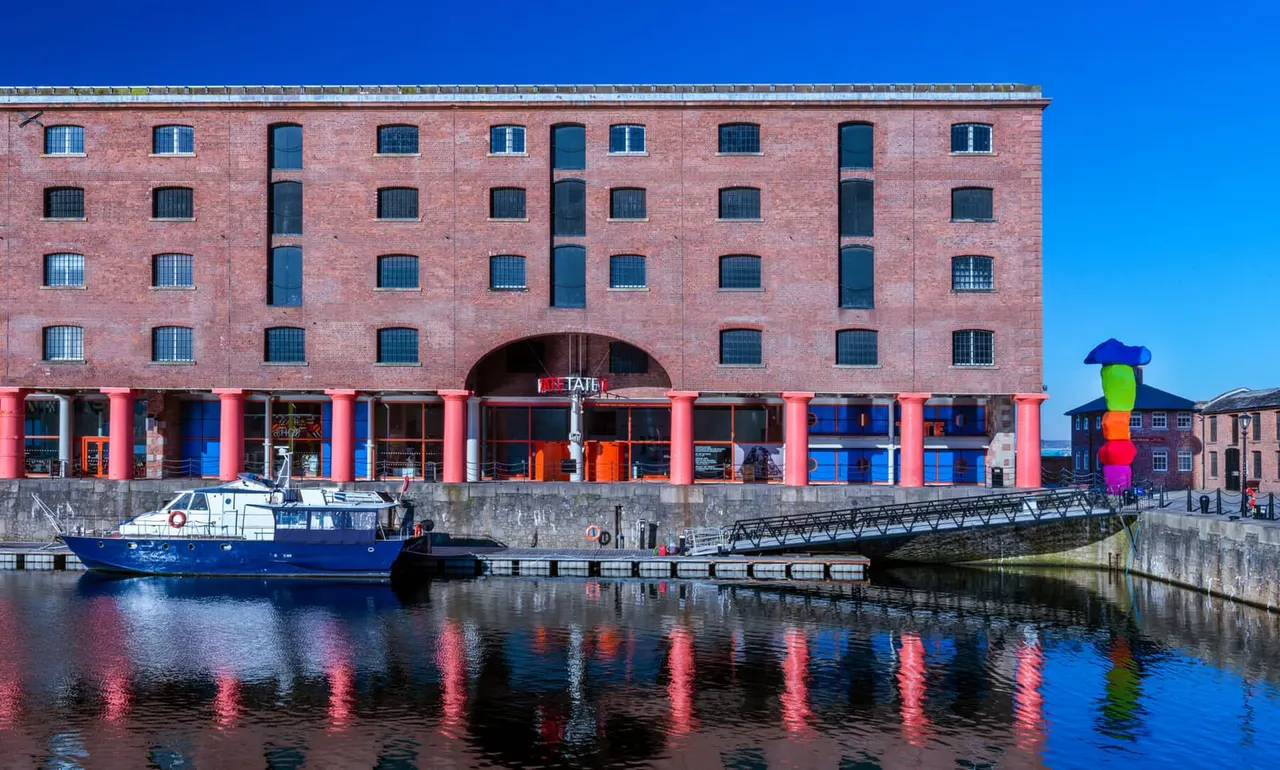 Red brick warehouse building with pink pillars at waterfront, blue boat moored beside dock.
