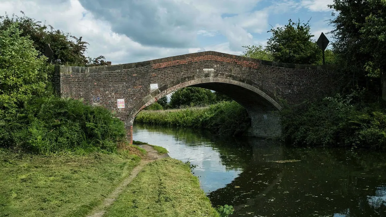 Historic brick arch bridge spanning a canal with towpath and vegetation on either side.