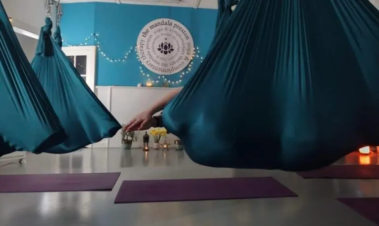 Two people in blue aerial yoga hammocks suspended in a studio with purple mats below.