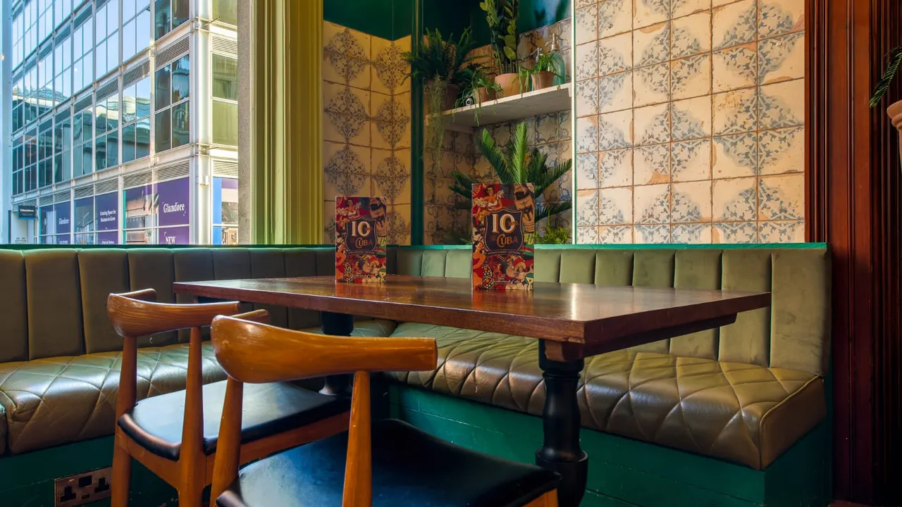 Interior dining booth with wooden table, mid-century chairs, green leather seating and potted plants beside city building views.