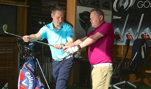 Two men examining a golf club together in a pro shop, with golf bags and equipment visible in the background.