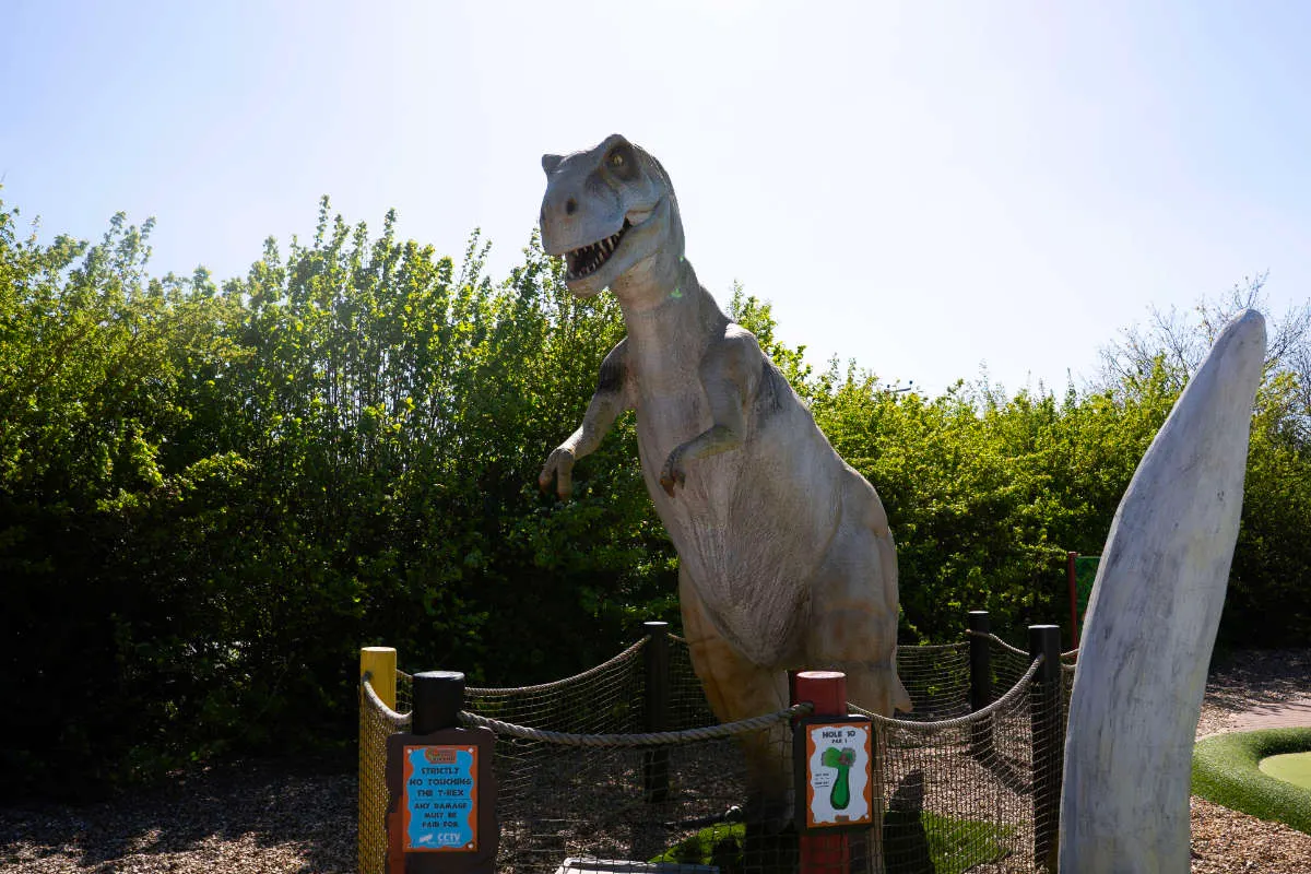 Large dinosaur sculptures displayed in a golf course attraction surrounded by green foliage and fencing.