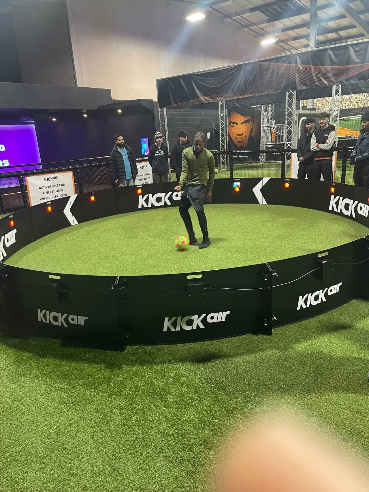 A person kicks a football on an indoor artificial pitch at an entertainment venue, with spectators watching from behind barriers.