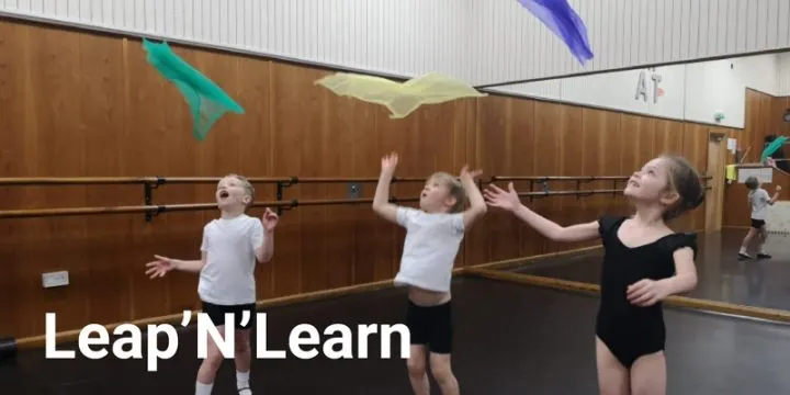 Children juggle and catch paper aeroplanes in a wooden-walled studio during a movement activity.