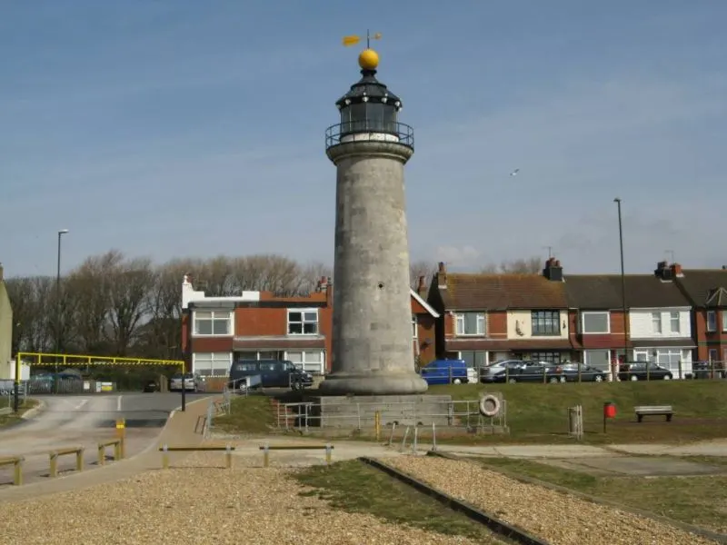 A stone lighthouse with a golden top stands on a shingle beach beside residential buildings under clear sky.