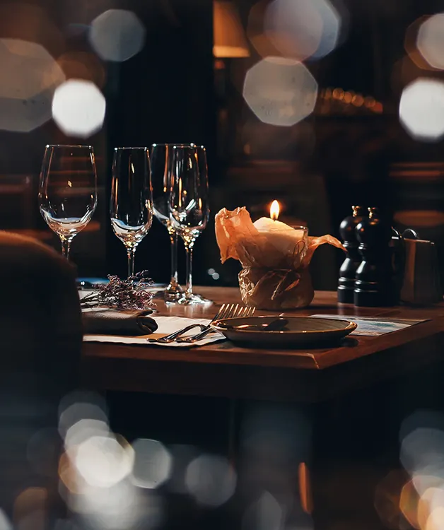 Elegantly set dining table with wine glasses, plate, cutlery, lit candle and blurred warm lighting in background.