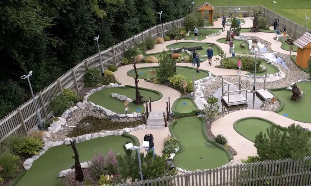 Aerial view of an outdoor mini-golf course with families playing, surrounded by landscaping, wooden fencing and trees.