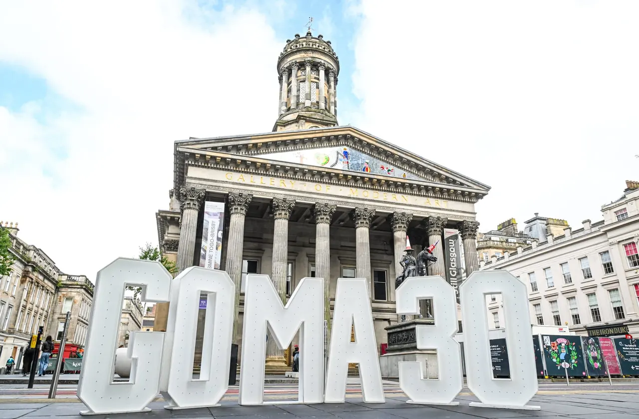 Historic neoclassical building with columned portico and domed tower, with large white "GoMA" letters in the foreground in an urban plaza.