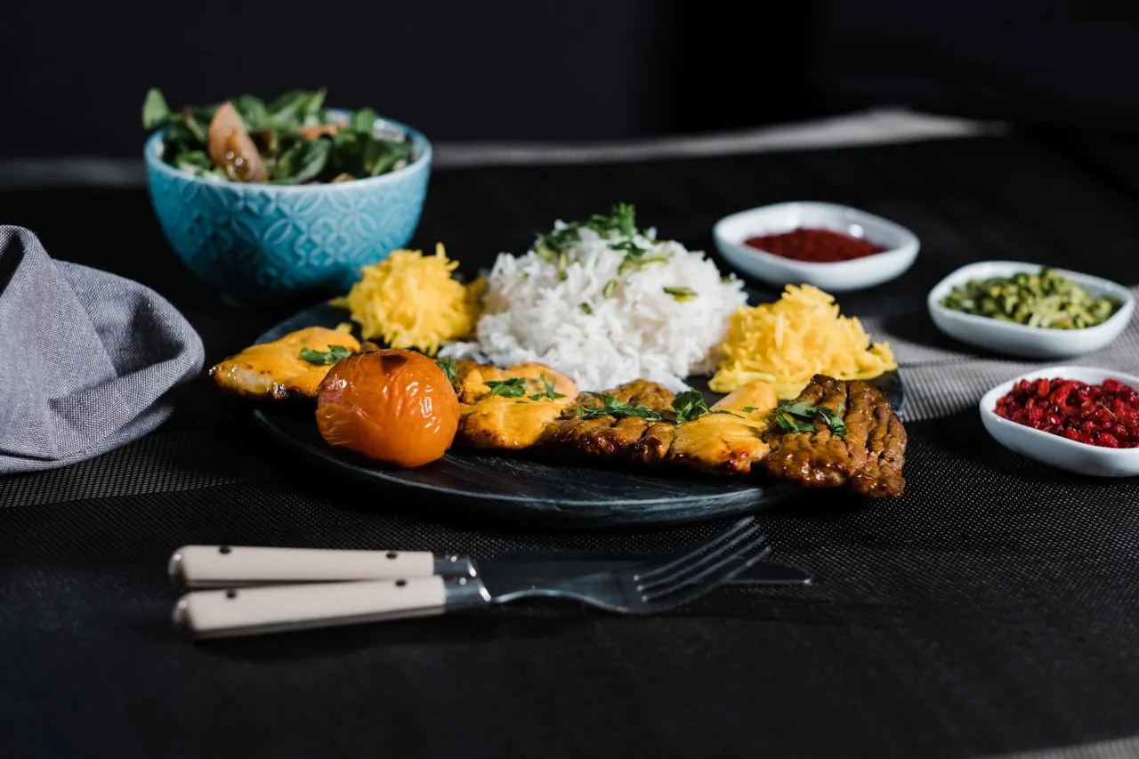 Grilled meat skewers with saffron rice, roasted vegetables and side condiments on a dark table setting.