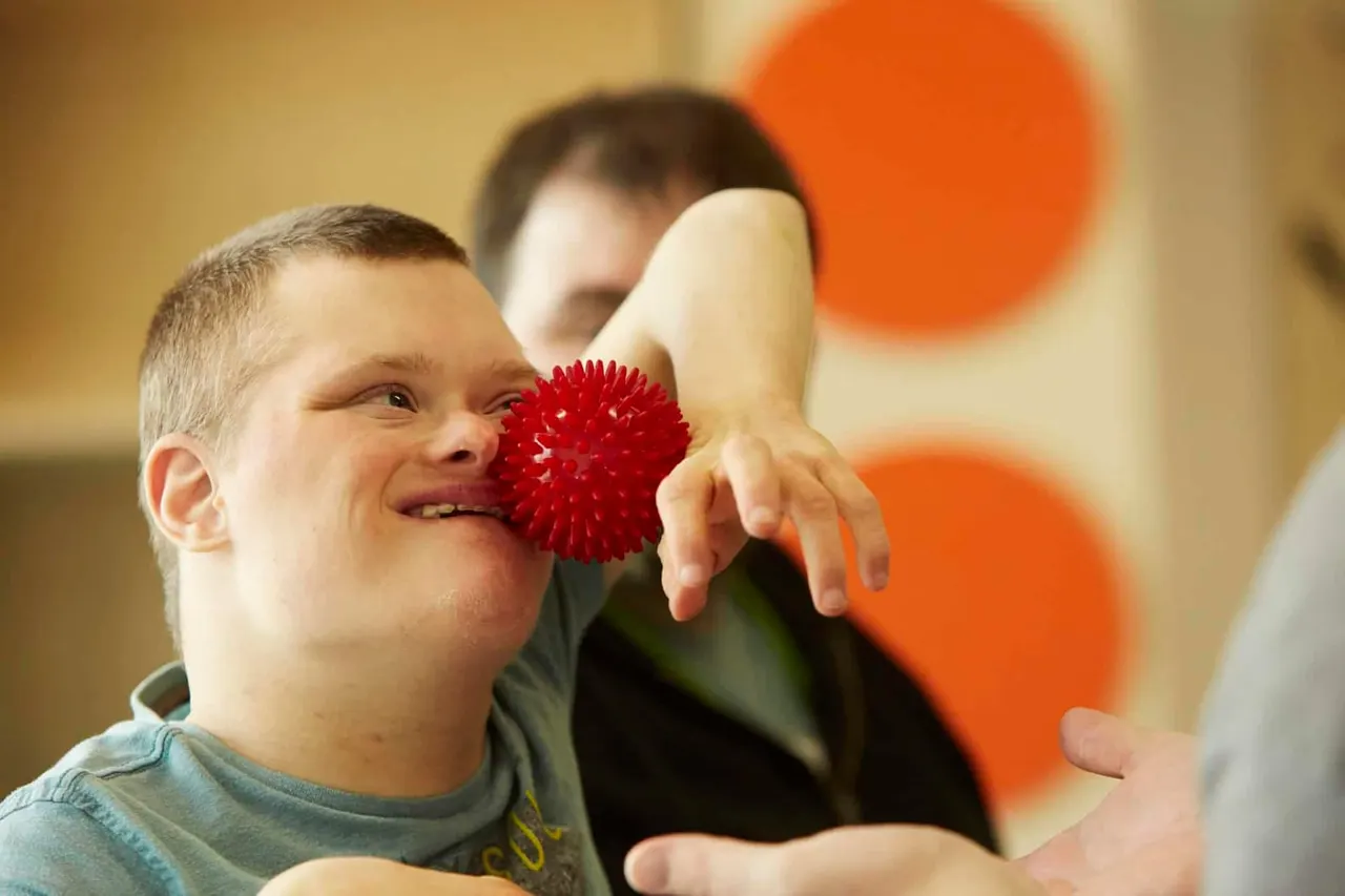 Young child holding a red textured ball near their face, smiling, with another child and adult nearby and orange balloons in the background.