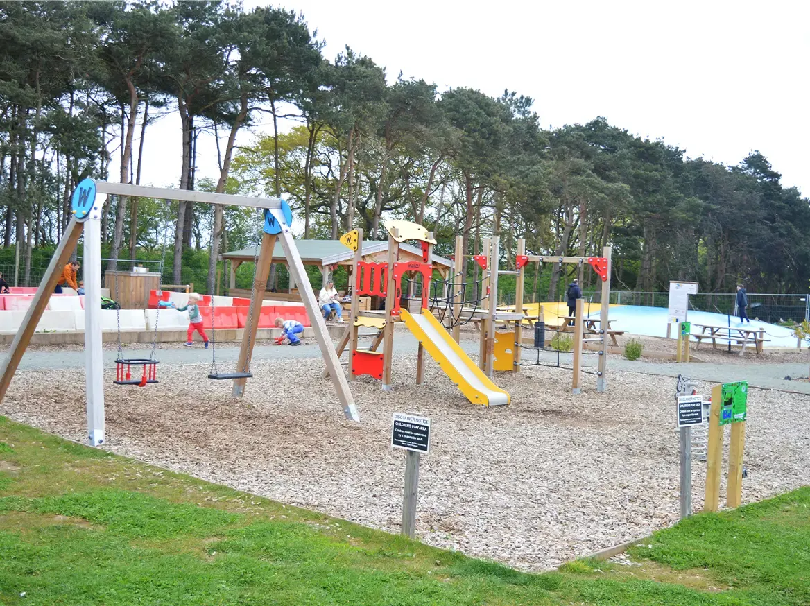 Children playing on a colourful wooden playground with swings, slides and climbing equipment surrounded by tall trees.