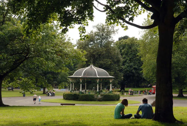 Two people sitting on grass beneath a large tree, with a domed bandstand and mature trees visible across the park.