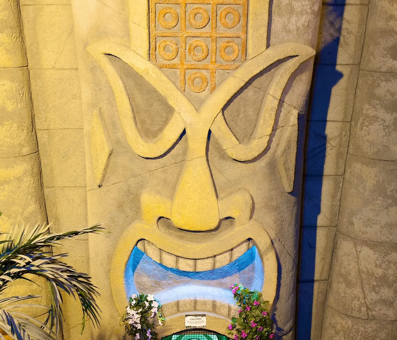 Stone carving of a smiling face with large ears and blue mouth, with flowering plants at its base.