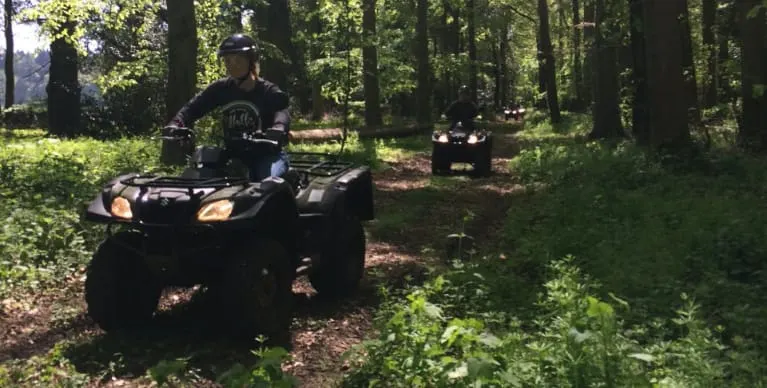 People riding quad bikes on a woodland trail lined with tall trees and green undergrowth.