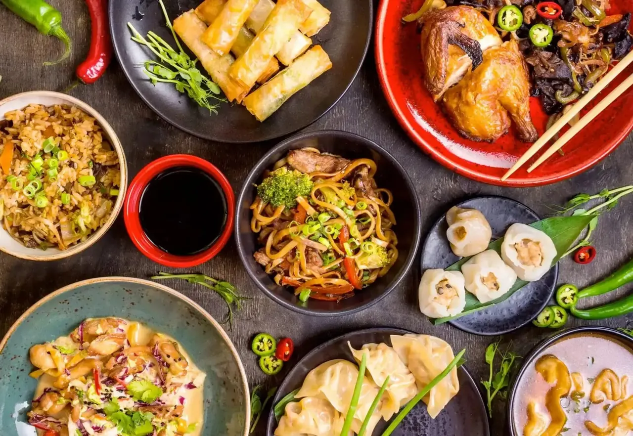Overhead view of Asian dishes including fried rice, spring rolls, noodles, dumplings and roasted poultry on colourful plates.