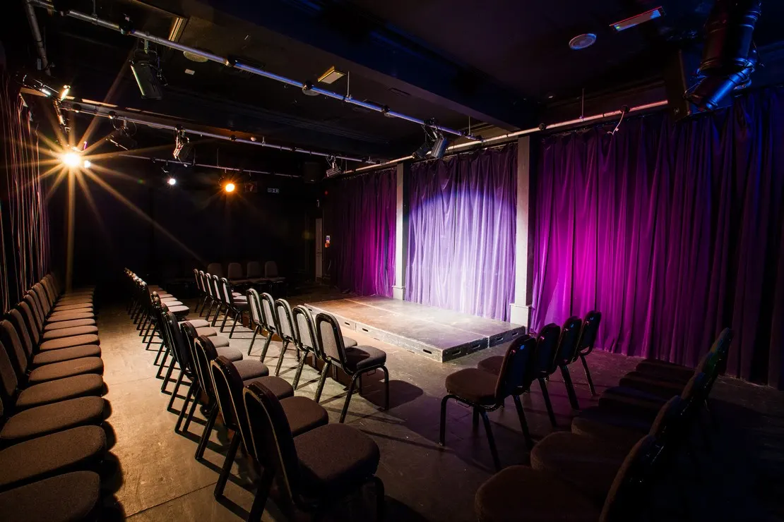 Empty theatre auditorium with rows of black chairs facing a lit stage with purple curtains and professional stage lighting.