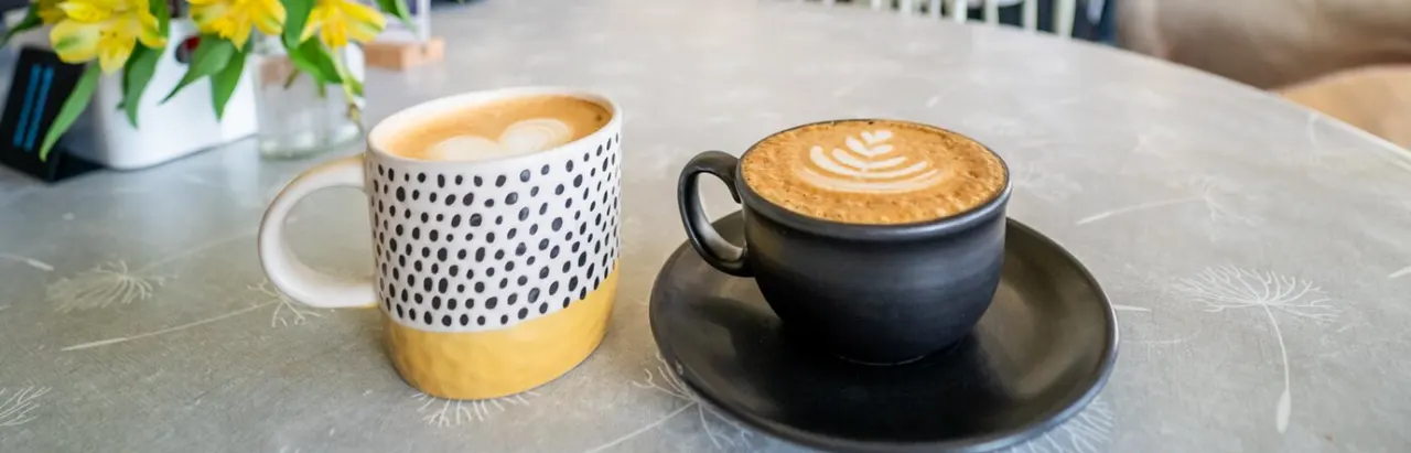 Two speciality coffees with latte art sit on a table next to yellow flowers in a vase.