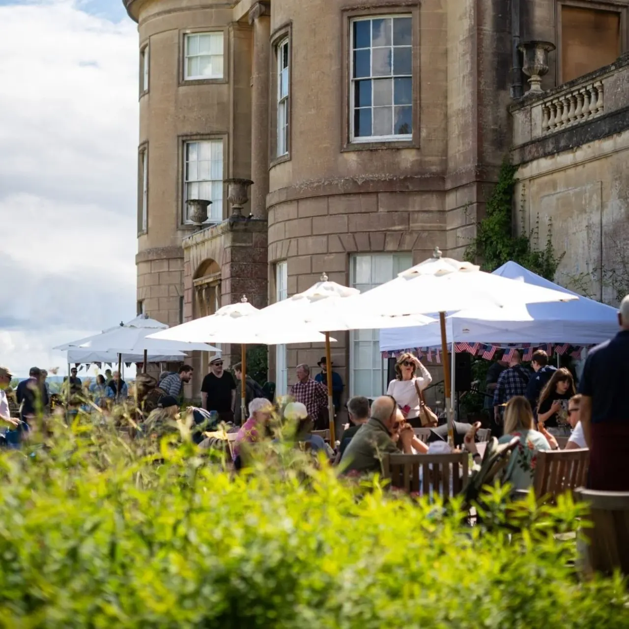 Guests socialising under white umbrellas on a terrace outside a historic stone mansion on a sunny day.