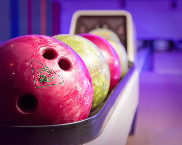 Colourful bowling balls lined up in a ball return, with pink ball in focus at a ten-pin bowling alley.